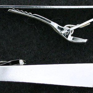 May include: Two silver-colored tie clips are displayed against a dark background. The top clip is a simple bar with a curved end and a hinged clip. The bottom clip is a rectangular bar with a hinged clip.