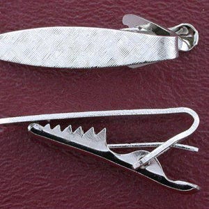 May include: Two silver-colored tie clips. One clip is oval with a textured pattern. The other clip is a bar style with a sawtooth edge. Both clips are made of shiny metal and are displayed against a burgundy background.