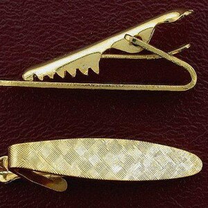 May include: Two gold-colored tie clips are displayed. The top clip has a sawtooth edge and a hinged design. The bottom clip is an oval shape with a textured, woven pattern. Both clips are shown against a dark, textured background.