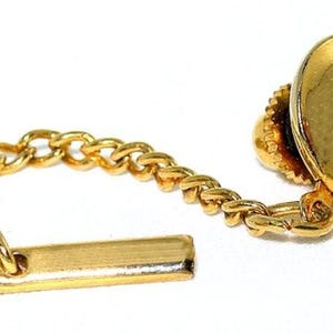 May include: A gold-toned tie tack featuring a vibrant red gemstone. The tie tack has a classic design with a decorative chain and a bar for securing it to the garment. The gemstone is round and set in a detailed, ornate setting.