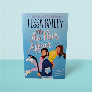 May include: A paperback novel titled "The Au Pair Affair" by Tessa Bailey. The cover art depicts a man and woman, with the title in a decorative font. The background is a light shade of blue.