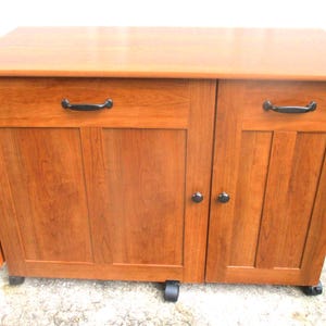 May include: A wooden cabinet with a warm, reddish-brown finish. It features two doors, a drawer, and black hardware. The cabinet is on wheels, and a side panel is folded down. The cabinet is likely used for storage.