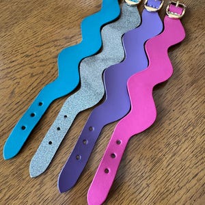 May include: Four wavy leather bracelets in turquoise, silver glitter, purple, and pink. Each bracelet has a gold-colored buckle and three holes for adjustment. The bracelets are displayed on a wooden surface.