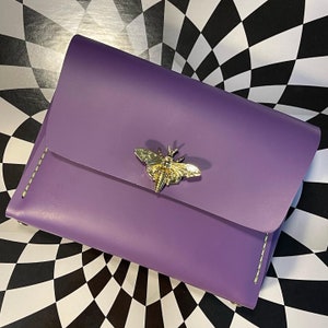 May include: A purple leather wallet with a gold butterfly design. The wallet has a flap closure and a stitched edge.