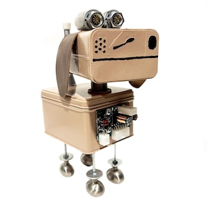 May include: A handmade robot dog sculpture constructed from recycled metal parts. The dog has a tan rectangular head with circular eyes, a brown leather strap, and a square body. The legs are made of silver spheres.
