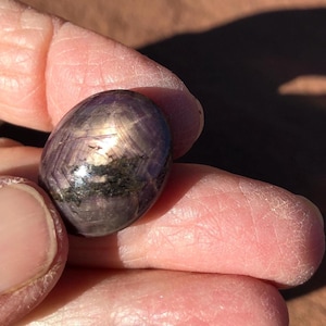 May include: A polished, oval-shaped cabochon gemstone featuring a deep purple and reddish-brown hue. The stone displays a distinct six-rayed star effect, known as asterism, with a bright, reflective light reflection across its smooth, rounded surface.