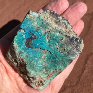 Könnte beinhalten: Ein grob behauener Stein mit einer lebendigen türkisen und schwarzen Farbpalette. Das Mineral hat eine strukturierte Oberfläche mit grünen und braunen Bereichen. Der Stein wird in einer Hand gehalten und zeigt seine natürliche Schönheit.