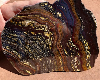 May include: A polished, irregularly shaped stone with a striking pattern of swirling bands in shades of brown, gold, red, and blue. The stone has a natural, raw edge and a polished surface, showcasing its unique mineral composition.