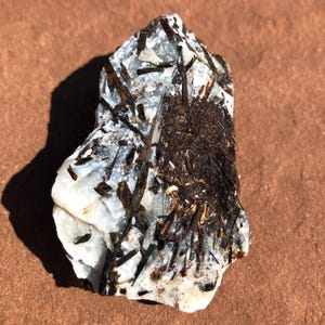May include: A raw mineral specimen featuring a white, translucent quartz matrix embedded with numerous dark brown to black, needle-like tourmaline crystals. A dense cluster of these dark, fibrous inclusions is concentrated on the right side.