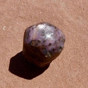 May include: A dark purple and silver gemstone with a rough, faceted surface. The gemstone is sitting on a red, sandy surface.