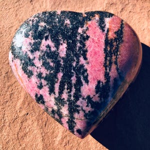 May include: A heart-shaped stone with a pink and black pattern. The pink areas have a marbled appearance, while the black sections are more solid. The stone is smooth and polished, with a natural, earthy aesthetic.