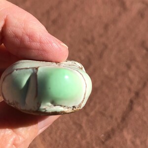May include: A small, rounded, ceramic object with a light green, glossy surface. The object has brown lines and a textured, off-white edge. The object is held in a hand against a brown background.