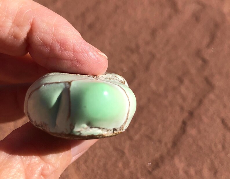 May include: A small, rounded, ceramic object with a light green, glossy surface. The object has brown lines and a textured, off-white edge. The object is held in a hand against a brown background.