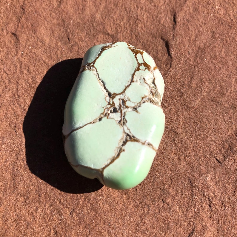 May include: A polished, oval-shaped gemstone with a pale green hue and a network of brown veins. The stone has a smooth, glossy surface and is set against a reddish-brown background. The gemstone is a natural, organic material.