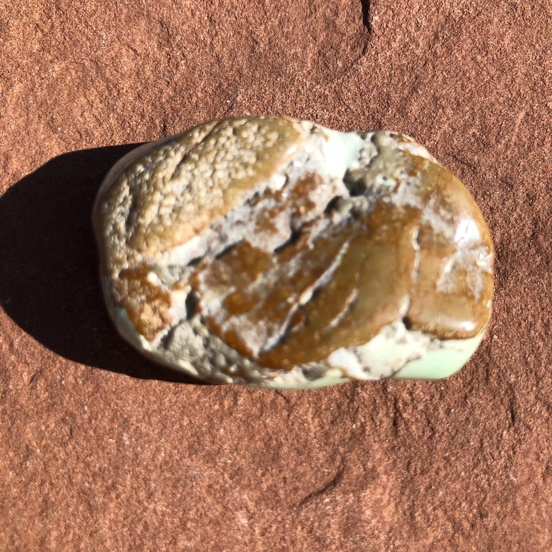 May include: A smooth, rounded stone with a mix of colors. The stone features areas of tan, brown, and light green. The surface has a textured appearance in some areas and a polished look in others. The stone is resting on a reddish-brown surface.