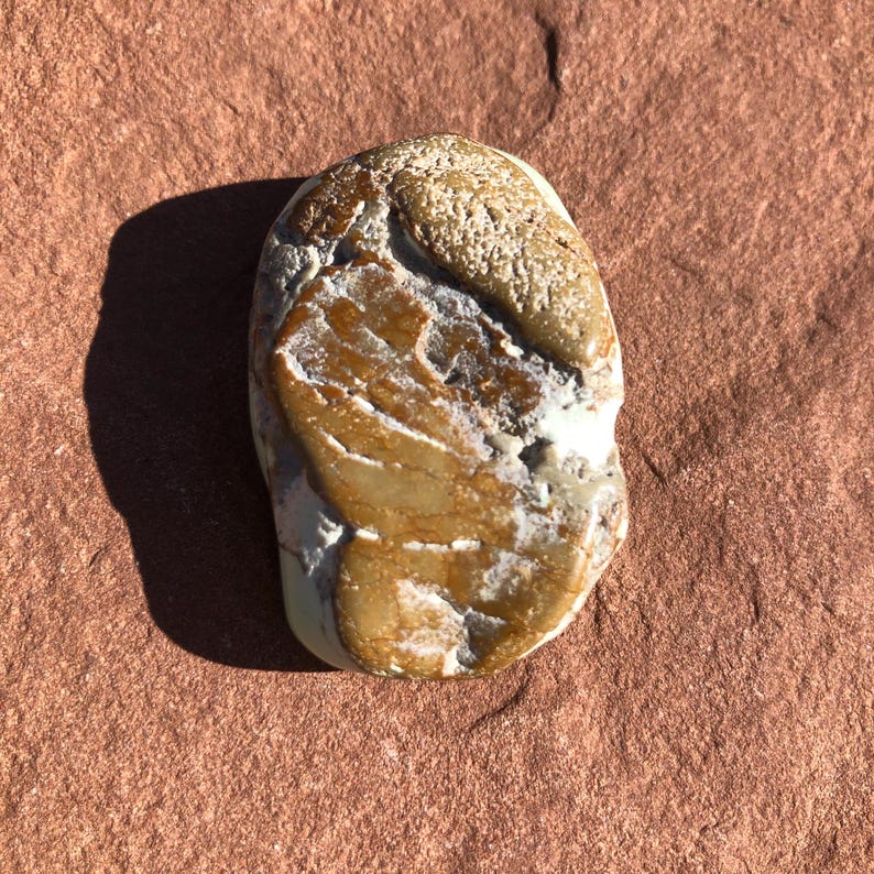 May include: A smooth, oblong stone with a mix of tan, brown, and white hues. The stone has a textured surface and a rounded shape, with a lighter-colored area at the top. The stone is set against a reddish-brown background.