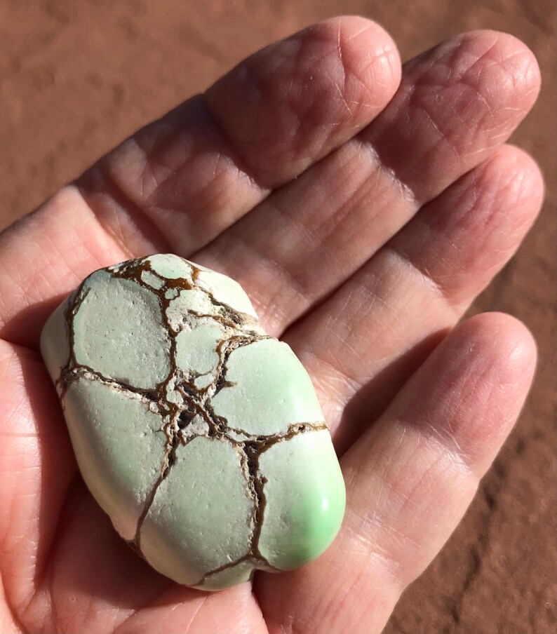 May include: A light green, oblong stone with brown veining. The stone has a smooth, polished surface on one end and a rough, textured surface on the other. The stone is held in a hand.