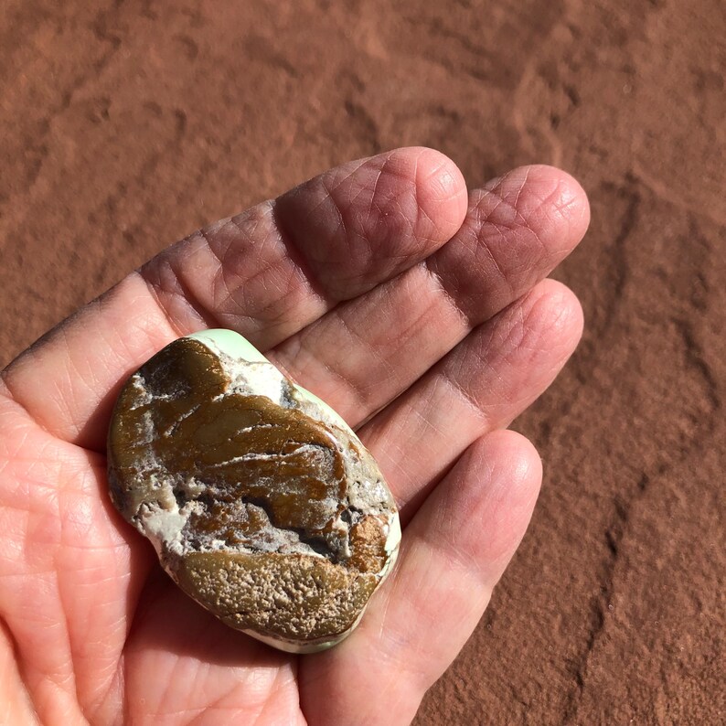 May include: A smooth, naturally shaped stone with a mix of colors. The stone has a light green area, a brown central section, and white and tan edges. The stone is held in an open hand, showcasing its natural texture and unique color variations.