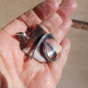 May include: A silver pendant with a large, rectangular, multi-colored stone. The stone has shades of pink, white, brown, and black. The silver is twisted and forms a loop at the top for a chain.