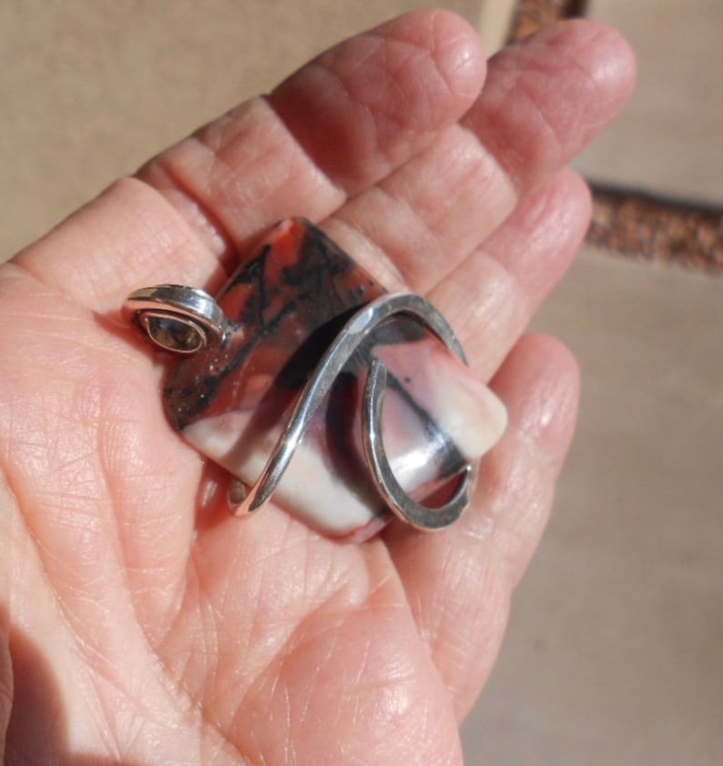 May include: A silver pendant with a large, rectangular, multi-colored stone. The stone has shades of pink, white, brown, and black. The silver is twisted and forms a loop at the top for a chain.