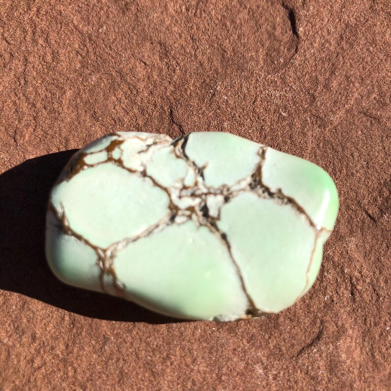 May include: A polished, light green gemstone with brown veining. The stone has a smooth, rounded shape and is set against a textured, brown background. The gemstone's color is a soft, pale green.