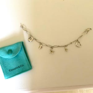 May include: Silver charm bracelet with various charms, including a horseshoe, star, and heart. A small, turquoise pouch with the Tiffany & Co. logo is next to the bracelet. The bracelet is on a white surface.