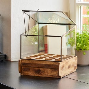 Può includere: Una scatola di legno con un coperchio di vetro e un coperchio incernierato. La scatola ha una griglia di scomparti all'interno. La scatola è realizzata in legno naturale e ha un aspetto rustico.