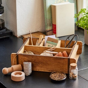 Könnte beinhalten: Eine Holzkiste mit Trennwänden, perfekt zum Organisieren von Gartenutensilien. Sie enthält Schnur, Saatgutpakete, kleine Werkzeuge und eine Gartenschere. Die Kiste steht auf einer schwarzen Oberfläche.