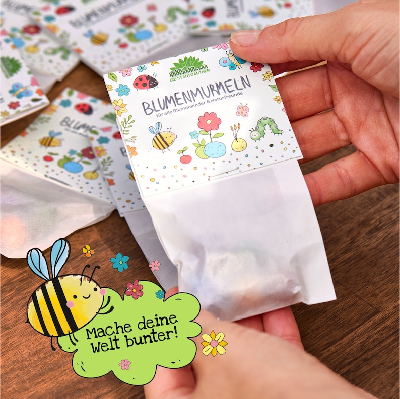 May include: A white paper bag with a colorful label that says "Blumenmurmeln" for all flower children and nature lovers. The label features a cartoon bee, flowers, and a caterpillar. The bag is held in a hand and has a small, colorful object inside. The text "Mache deine Welt bunter!" is written on a green speech bubble with a cartoon bee.