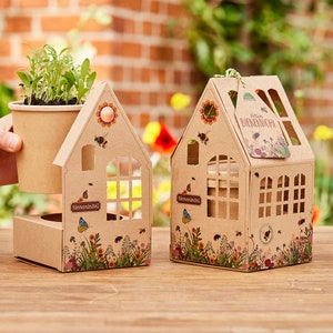Op de afbeelding: Twee bruine kartonnen huizen met uitgesneden ramen en bij-thema decoraties. De huizen zijn ontworpen om kleine potplanten te bevatten. De tekst "Bienen-Sel" is gedrukt op de voorkant van elk huis. De tekst "Ratum BIENENVOLL" is gedrukt op het rechter huis.