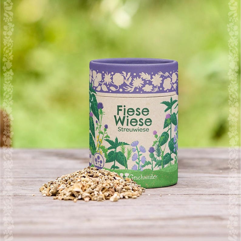 Puede incluir: Un recipiente cil&iacute;ndrico con el texto "Fiese Wiese Streuwiese" e ilustraciones bot&aacute;nicas. El recipiente es morado en la parte superior y verde en la inferior, con una secci&oacute;n central beige. Un mont&oacute;n de hierbas secas est&aacute; delante del recipiente.
