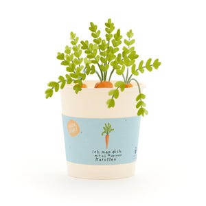 Könnte beinhalten: Ein verspielter Pappbecher mit einem hellblauen Band mit einer Karottenillustration und deutschem Text. Der Becher enthält drei orangefarbene Karotten mit grünen Blattoberteilen, wodurch ein verspieltes, gartenorientiertes Design entsteht.