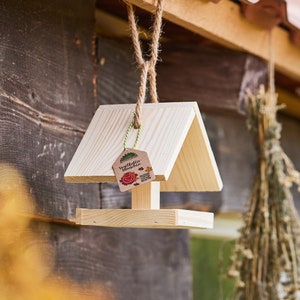 Op de afbeelding: Een houten vogelvoederhuisje met een klein dak en een bruin papieren label met de tekst "Vogelfutter Häuschen" met een rood hart en bladeren. Het huisje hangt aan een touw.