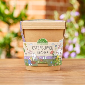 Può includere: Un vaso di carta marrone con coperchio, avvolto da una fascia decorativa con coniglietti e fiori dei cartoni animati. Il testo "OSTERBLUMEN BECHER" è stampato sulla fascia. Il vaso è appoggiato su una superficie di legno.
