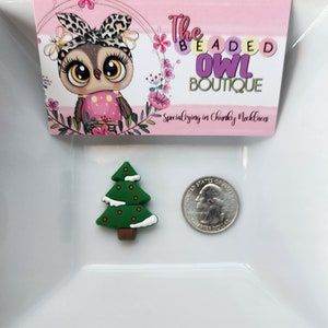 May include: A green Christmas tree charm with red dot accents and white snow-like trim. A U.S. quarter coin is next to the charm for size comparison. The image also includes a pink business card with an owl graphic and the text "The Beaded Owl Boutique".