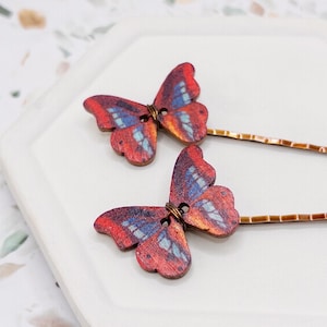 May include: Two wooden butterfly hair pins. The butterflies are painted with red, blue, and orange hues. The wings are detailed with a wood grain pattern. The pins are made of copper-colored metal.