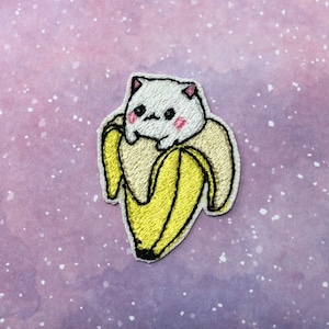 May include: White embroidered cat with pink cheeks sitting inside a yellow banana peel. The cat is looking to the left. The banana peel is embroidered with yellow and brown thread.