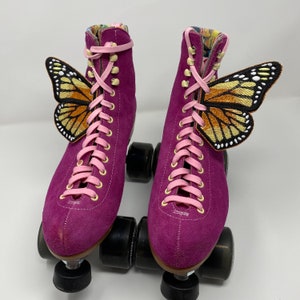 May include: A pair of purple suede roller skates with pink laces and butterfly appliques. The skates have black wheels and a pink and white floral pattern on the inside of the boot.