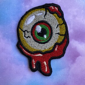 May include: A colourful embroidered patch of a cartoon eyeball with a red, green and white iris. The eyeball is dripping with red and black paint.