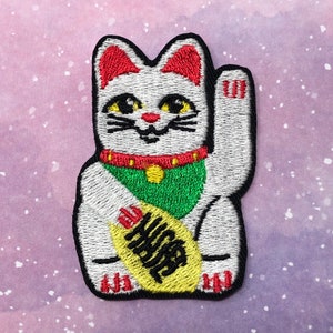 May include: White embroidered lucky cat with a red collar, green scarf, and a yellow coin with black kanji characters. The cat is waving its right paw.