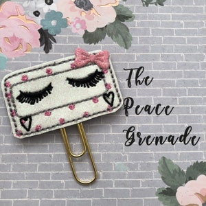 May include: A decorative paper clip featuring a white rectangle with black eyelashes, a pink bow, and small pink polka dots. The text "The Peace Grenade" is written in black script. The clip is gold-colored.