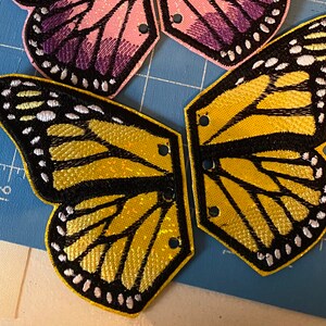 May include: Two yellow and black butterfly wings with white accents. The wings are made of fabric and have a shiny finish. They are attached to each other with a small piece of fabric.