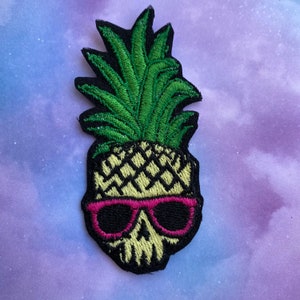 May include: Embroidered patch featuring a pineapple skull design. The patch has a black outline, with a green pineapple top and a skull face wearing pink sunglasses. The skull is a light yellow color.
