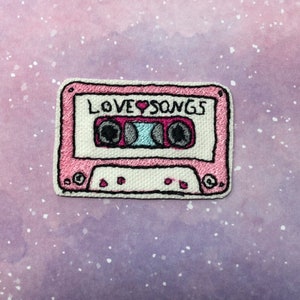 May include: Pink embroidered cassette tape patch with the text "LOVE SONGS" on it.