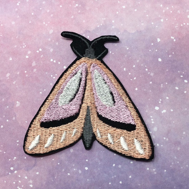 K&ouml;nnte beinhalten: Ein gestickter Motten-Patch in Rosa und Braun mit schwarzen Akzenten. Die Motte hat eine Fl&uuml;gelspannweite von etwa 5 cm.