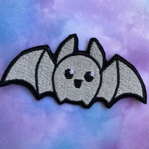 May include: A gray embroidered iron-on patch of a cartoon bat with big eyes and a smile. The bat has black outlines and white eyes.