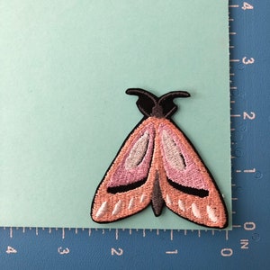 K&ouml;nnte beinhalten: Rosa-wei&szlig;er gestickter Motten-Patch mit schwarzen Akzenten. Die Motte ist etwa 5 cm breit.