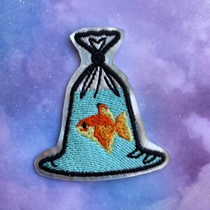 May include: A silver-colored embroidered patch of a plastic bag with a goldfish inside. The bag is tied at the top with a black string. The goldfish is orange and has black eyes.