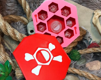 May include: A pink hexagonal dice tray with a skull and crossbones design on the lid. The tray has compartments for holding dice and a small compartment for a dice rolling tower. The lid is red with a white skull and crossbones design.