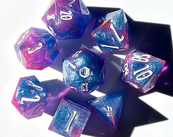 Puede incluir: Un conjunto de siete dados poliédricos de varias formas. Los dados son una mezcla de azul, rosa y morado con números blancos. Diseñados para juegos de rol de mesa.